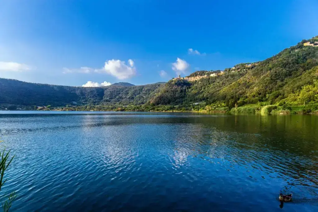 Italy Moto Tours Scenic view of Lake in Lazio, Italy, surrounded by forested hills and hilltop villages under a bright blue sky with soft clouds.