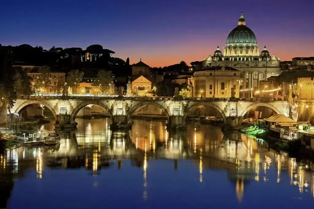 Italy Moto Tours Evening view of St. Peter’s Basilica and Ponte Sant'Angelo in Rome, Italy, with golden lights reflecting over the Tiber River.