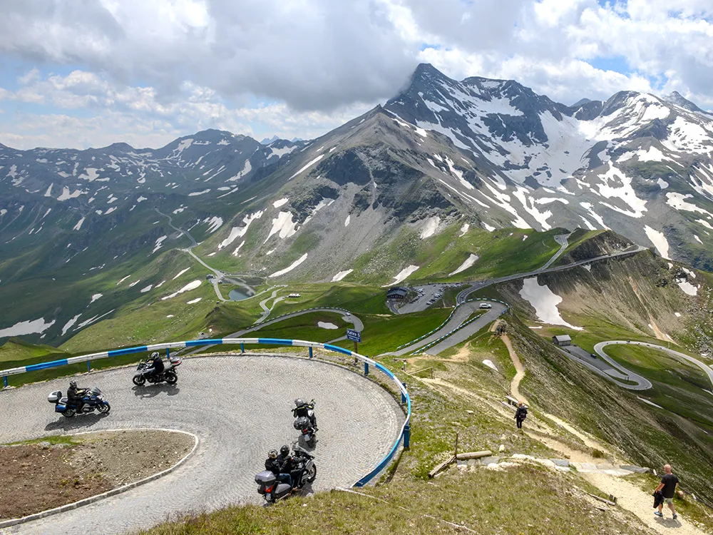 Guided motorcycle group negotiates a tight cobblestone hairpin on a high Dolomites alpine pass, with snow-capped peaks in the background