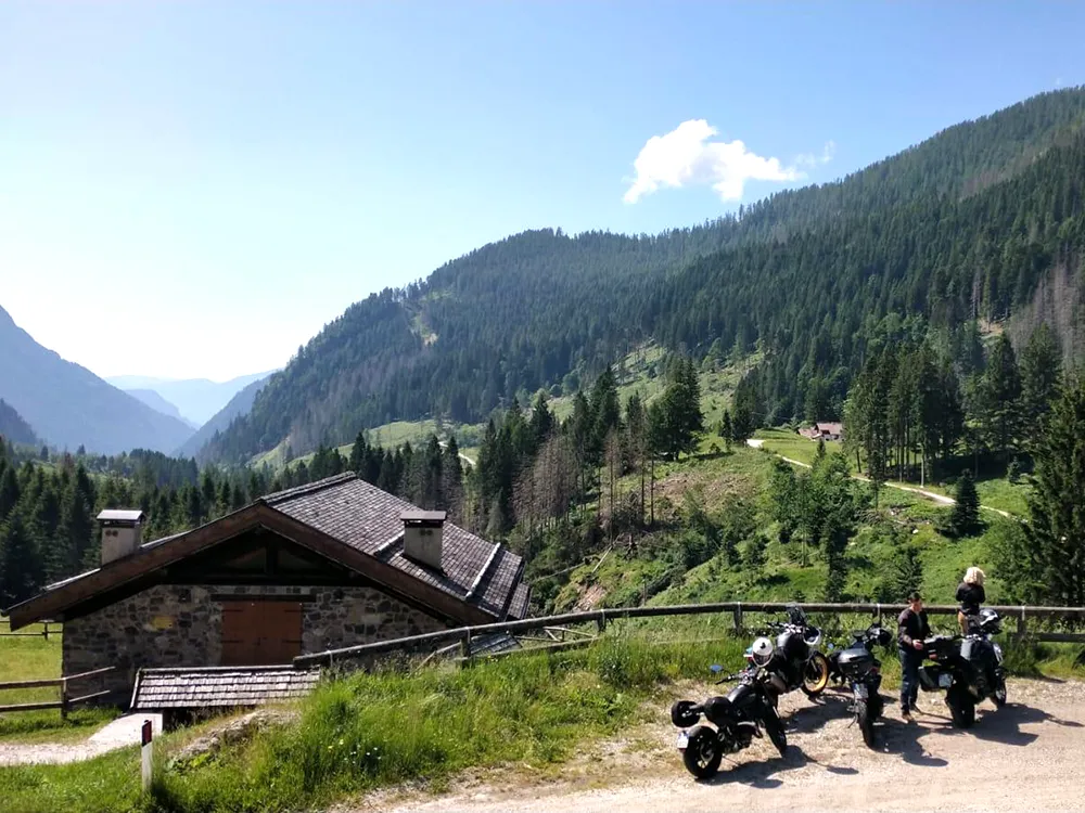 Italy MotoTours riders and motorcycles pause beside an alpine stone hut overlooking a lush Dolomites valley during a guided tour break
