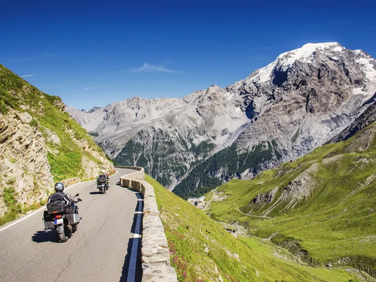 Horizontal image of Passo di Falzarego showing two motorcyclists riding along a winding mountain road framed by green slopes and towering snow‑capped peaks beneath a clear blue sky
