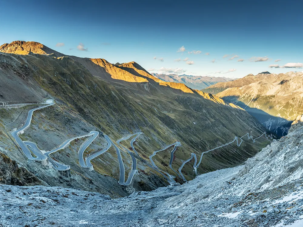 Horizontal panoramic view of the Stelvio Pass showing its dramatic hairpin bends and switchbacks climbing the rugged Italian Alps in evening light