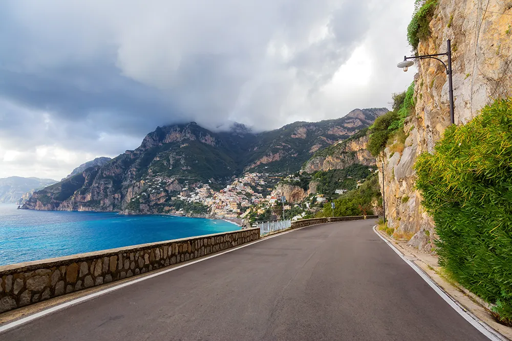 Italy Moto Tours Cliffside road hugs rock wall above the Tyrrhenian Sea, curving toward Positano on Italy's Amalfi Coast under dramatic clouds.