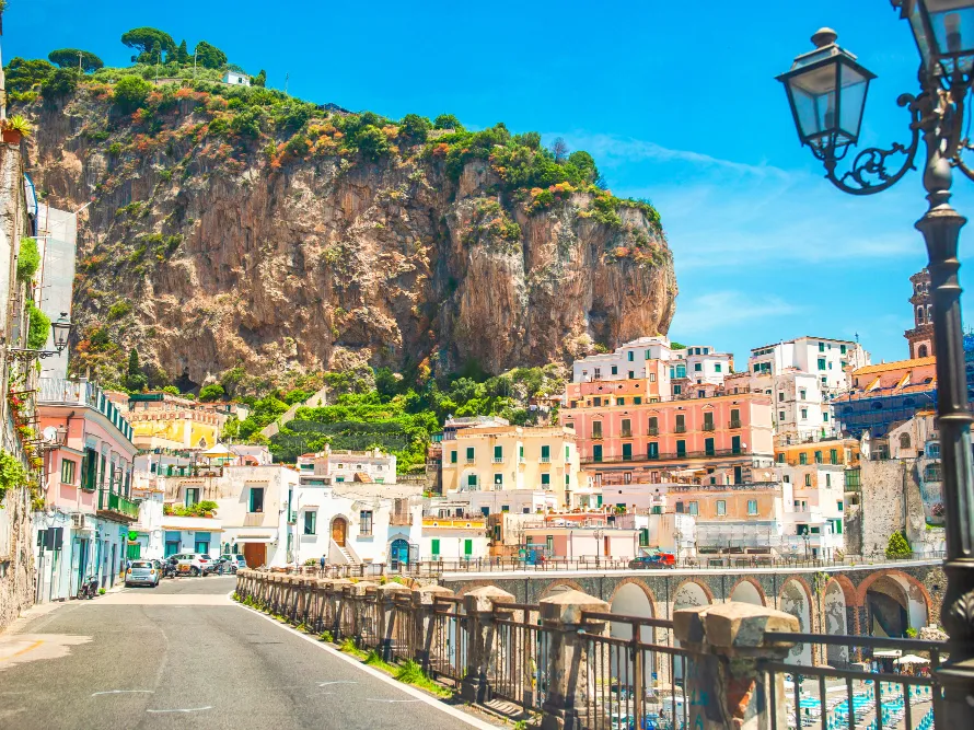 Italy Moto Tours Vibrant houses in the town of Amalfi, Italy, lining a quiet street with steep rocky cliffs in the background under a bright blue sky