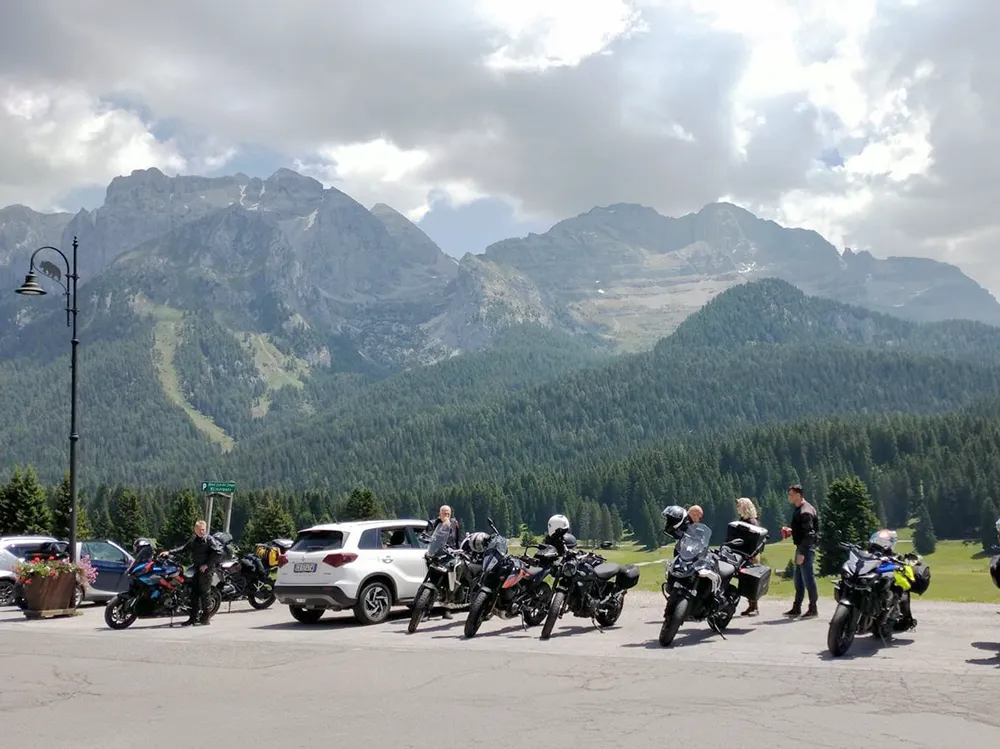 Lineup of touring motorcycles and support SUV parked near Dolomites mountains, riders chatting below rugged peaks