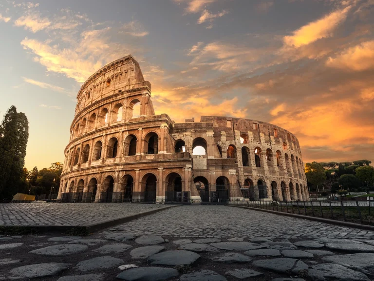 The Colosseum bathed in warm sunset light with the uneven cobblestone pavement of an ancient Roman road in front