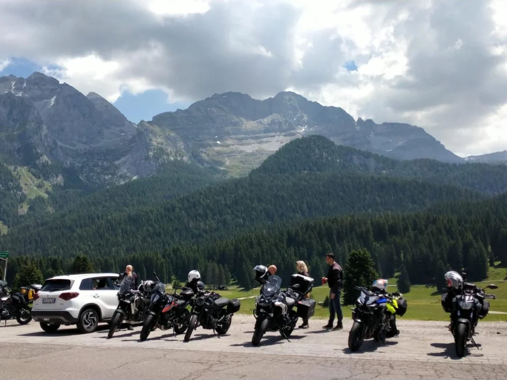 Italy Moto Tours Riders and parked motorcycles at an alpine viewpoint in Italy, with pine forests and rugged peaks in the background.