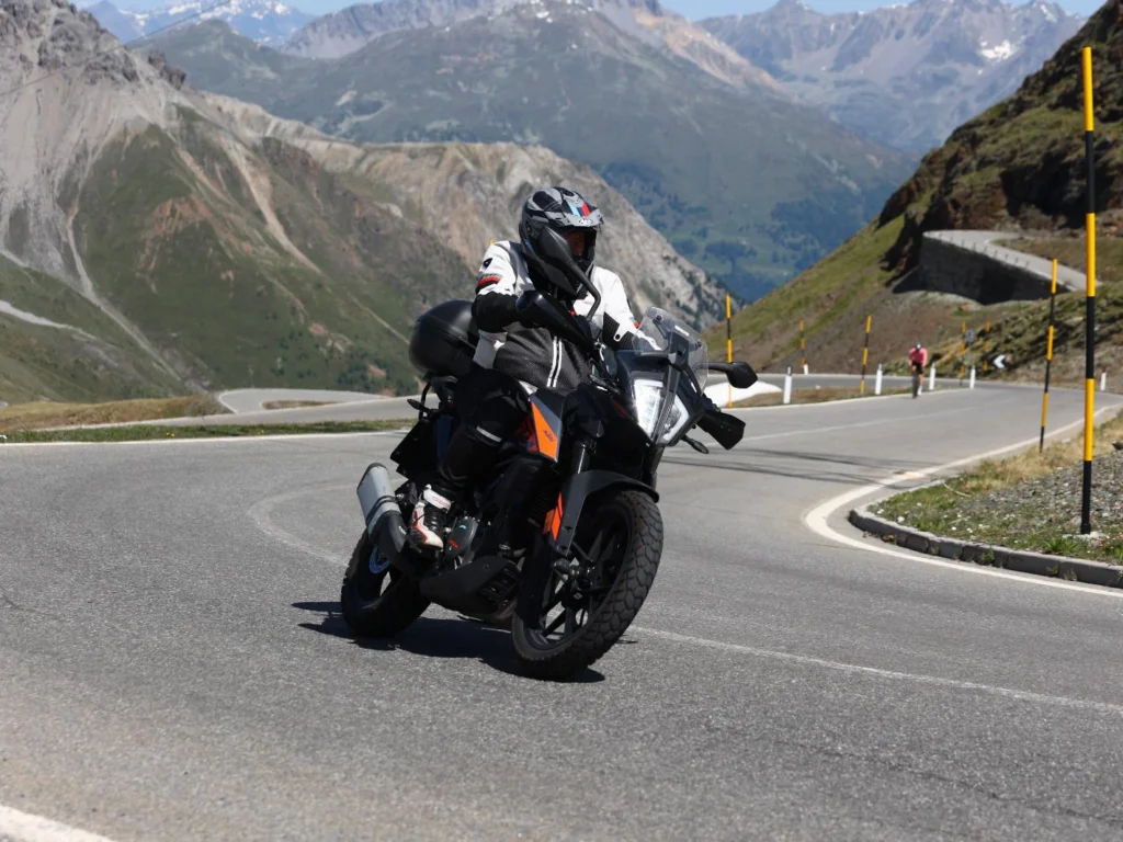 Italy Moto Tours Close view of a gloved rider controlling the bike mid-turn on a curving Alpine roadway marked with yellow posts, dramatic peaks all around.