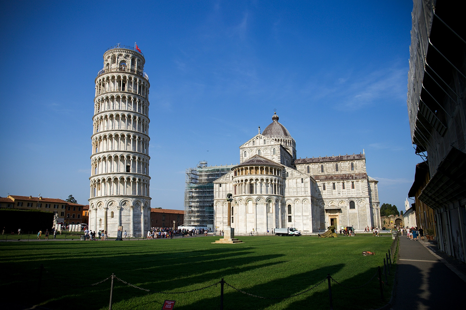 The Leaning Tower of Pisa beside Pisa Cathedral on a sunny day, with tourists gathering on the green lawn