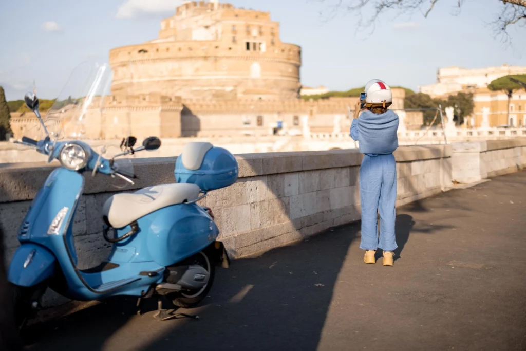 Italy Moto Tours Blue Vespa parked by the riverbank as a woman photographs Castel Sant’Angelo across the Tiber on a sunny day