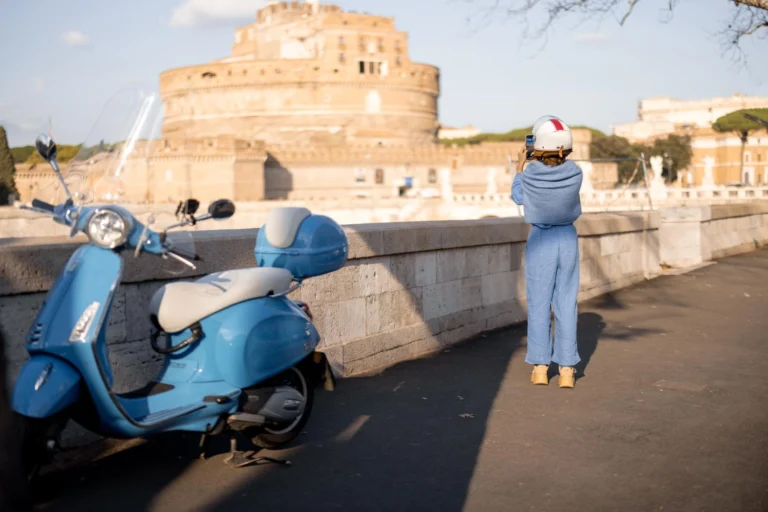 Blue Vespa parked by the riverbank as a woman photographs Castel Sant’Angelo across the Tiber on a sunny day