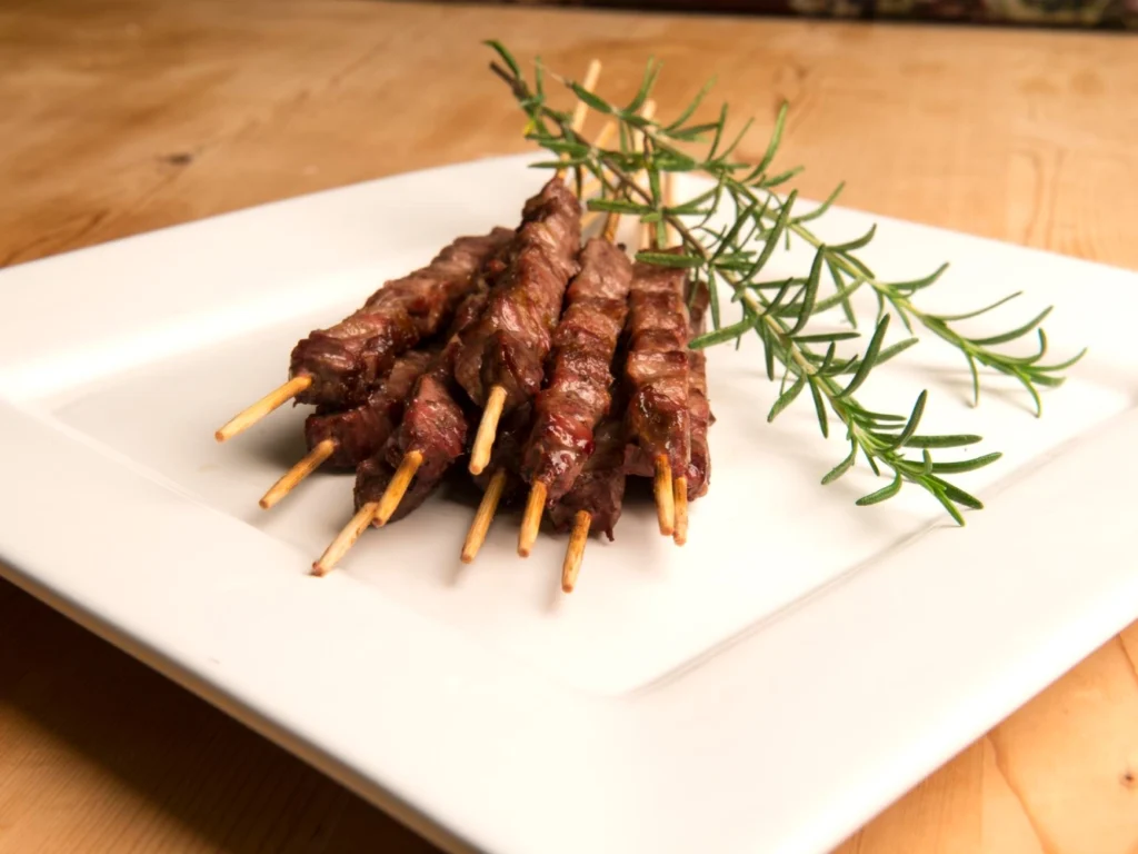 Italy Moto Tours Grilled lamb arrosticini skewers arranged on a white plate with a sprig of rosemary.