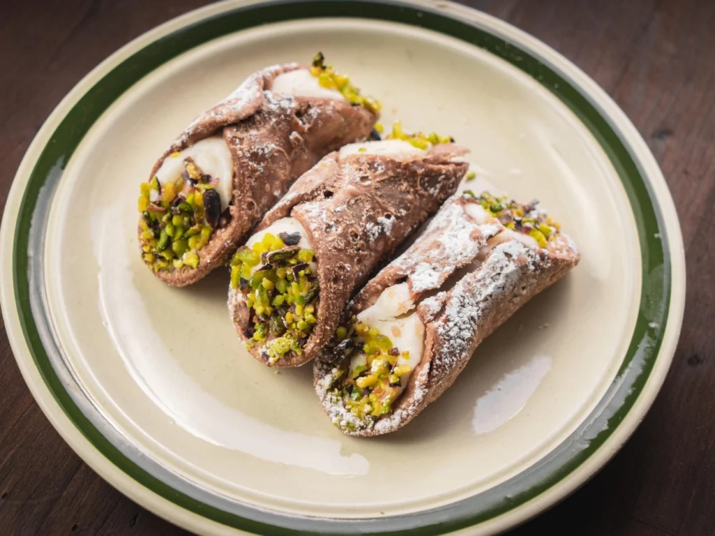 Italy Moto Tours Three Sicilian cannoli filled with sweet ricotta, pistachio-dipped ends and powdered sugar on a plate.