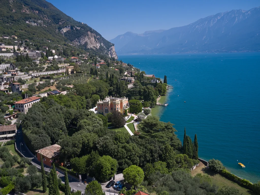 Italy Moto Tours Lake Garda’s western shoreline with villas, cypress trees, and mountains dropping to vivid blue water.
