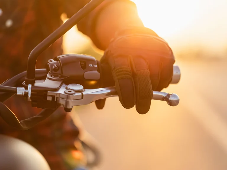 Italy Moto Tours Motorcyclist’s gloved hand resting on the brake lever of a handlebar, backlit by a warm golden sunset.