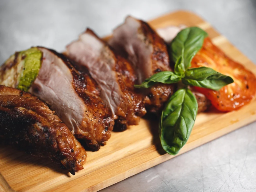 Italy Moto Tours Close-up of roasted pork slices served on a board, garnished with fresh basil and tomato, typical of Sardinia.