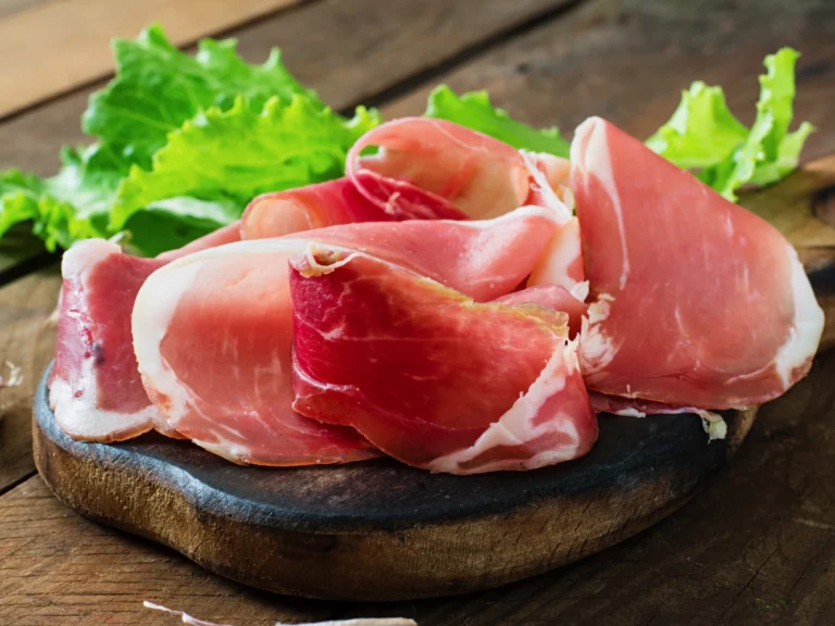 Italy Moto Tours Thin slices of Italian prosciutto crudo arranged on a small wooden board with fresh lettuce in the background.