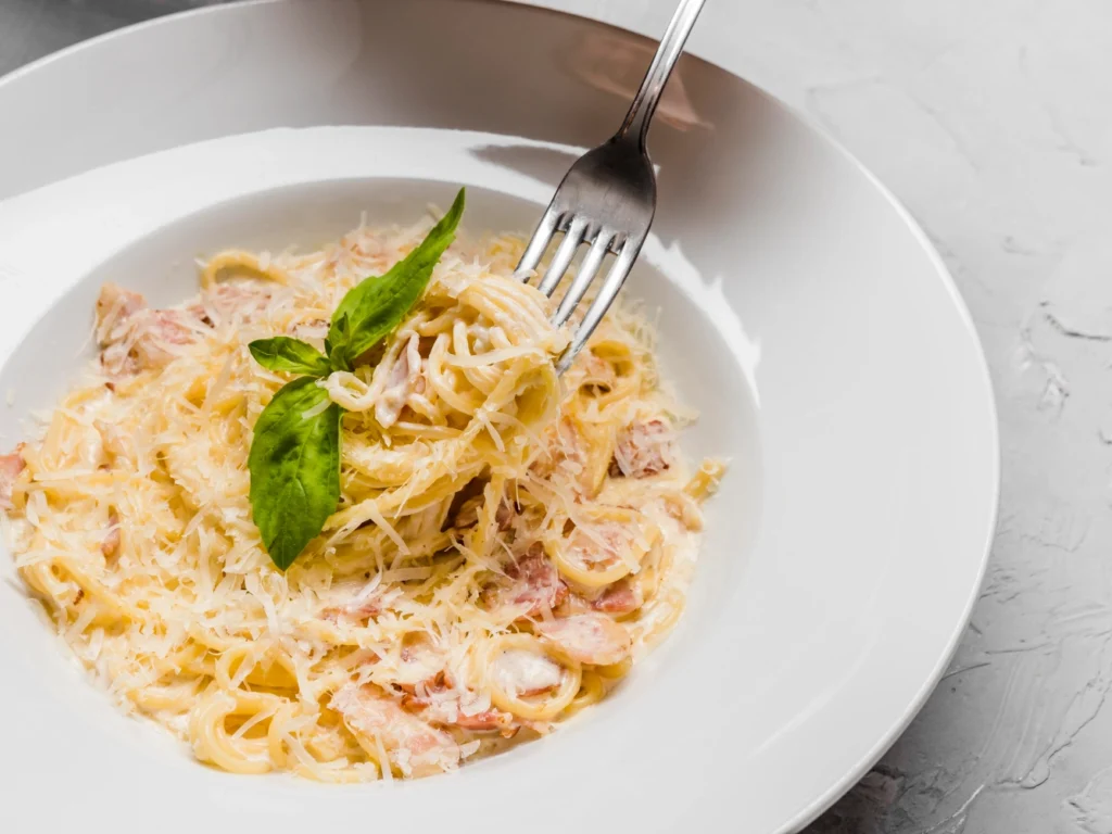 Italy Moto Tours Close-up of spaghetti twirled on a fork in a creamy cheese sauce with pancetta pieces and a basil garnish.