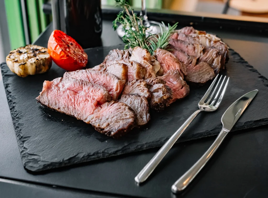 Italy Moto Tours Medium-rare sliced beef steak on a black slate with rosemary sprigs, grilled tomato and garlic, fork and knife beside it.