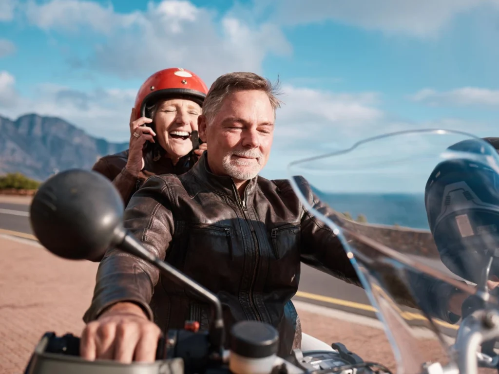 Smiling passenger adjusting a red helmet behind the rider on a motorcycle; both in brown leather jackets, cruising a coastal road with sea and mountains.