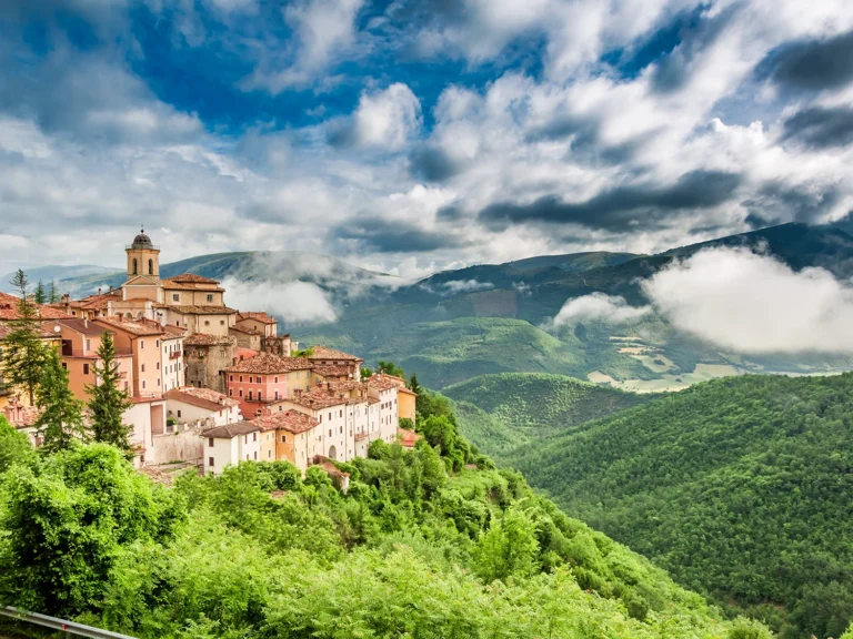 Italy Moto Tours Colorful medieval hill town perched on a ridge with forested mountains and low clouds, Umbria, Italy