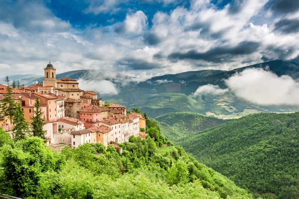 Italy Moto Tours Colorful medieval hill town perched on a ridge with forested mountains and low clouds, Umbria, Italy