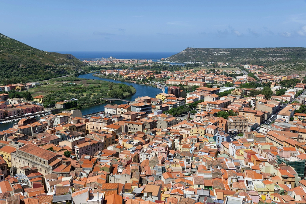 Italy Moto Tours Panoramic view over Bosa’s terracotta rooftops with the Temo River winding toward the Mediterranean between green hills.