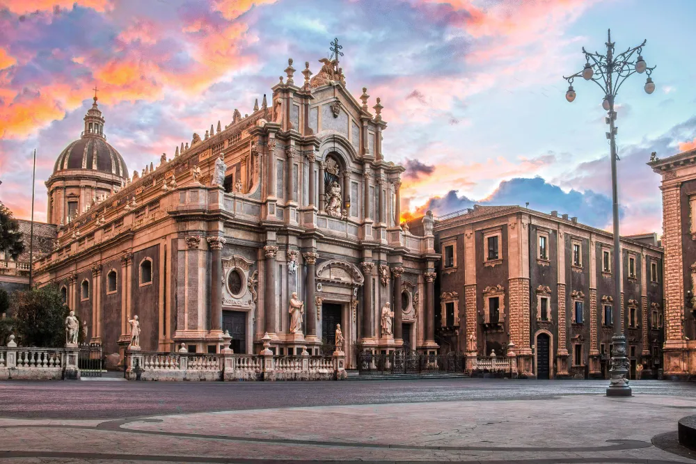 Italy Moto Tours Faсade of Catania’s cathedral on Piazza Duomo under a pink and blue evening sky.