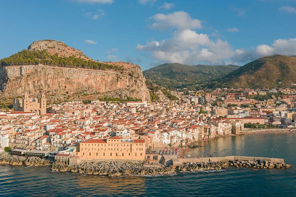 Italy Moto Tours Aerial view of Cefalù’s medieval center backed by La Rocca cliff and the Tyrrhenian Sea.