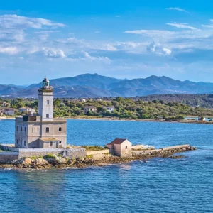Historic lighthouse on a tiny islet linked by a breakwater at the entrance to Olbia’s gulf, with hills and coastal homes in the background.