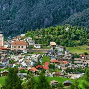 Tyrolean village with a tall church steeple, stone houses and green meadows at the base of forested mountains.