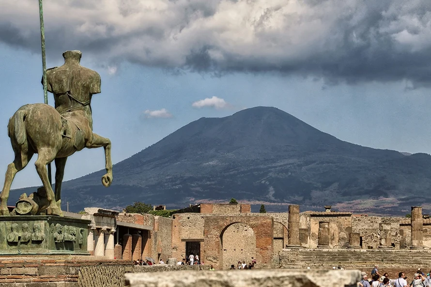 Italy Moto Tours Bronze centaur statue overlooking the ruins of Pompeii’s Forum, with Mount Vesuvius rising under dramatic clouds and visitors among ancient columns.