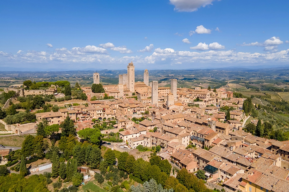 Italy Moto Tours Aerial panorama of San Gimignano in Tuscany, Italy, showing its medieval towers above terracotta rooftops and rolling countryside.