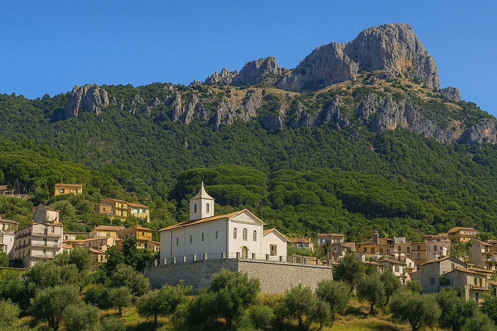 Italy Moto Tours White hilltop church above a colorful Sardinian village with pine-covered slopes and rugged limestone cliffs of the Supramonte massif.