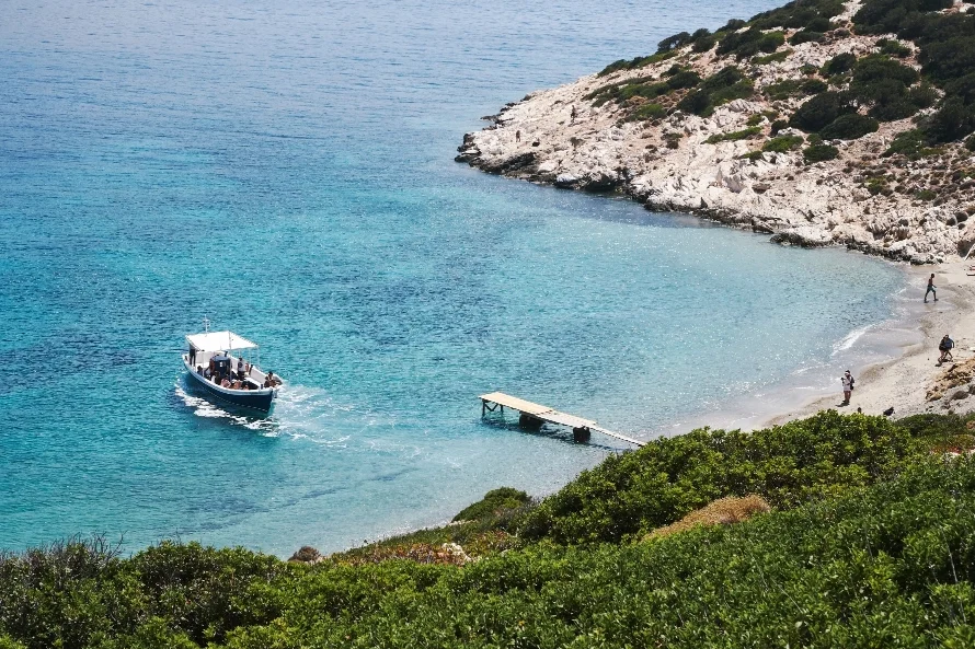 Italy Moto Tours Small tour boat approaching a tiny wooden jetty in a turquoise cove framed by low rocky headlands and scrub on Sardinia’s coast.