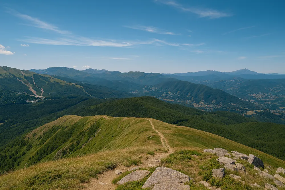 Italy Moto Tours Panoramic view from Abetone Pass with a grassy ridge trail, forested slopes, and distant Apennine ranges under a clear blue sky.