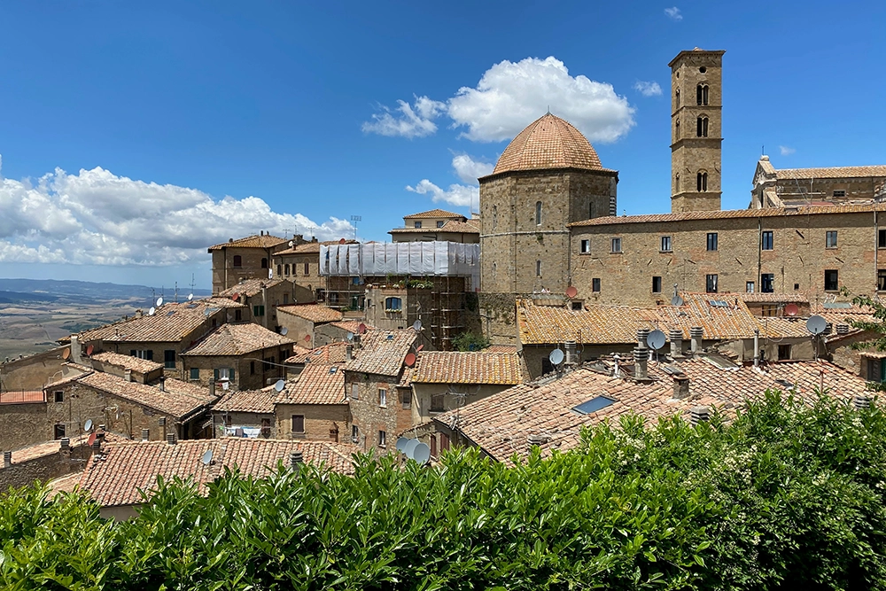 Italy Moto Tours Rooftop view of Volterra, Tuscany, with stone houses, the cathedral dome and a medieval bell tower under a blue sky.