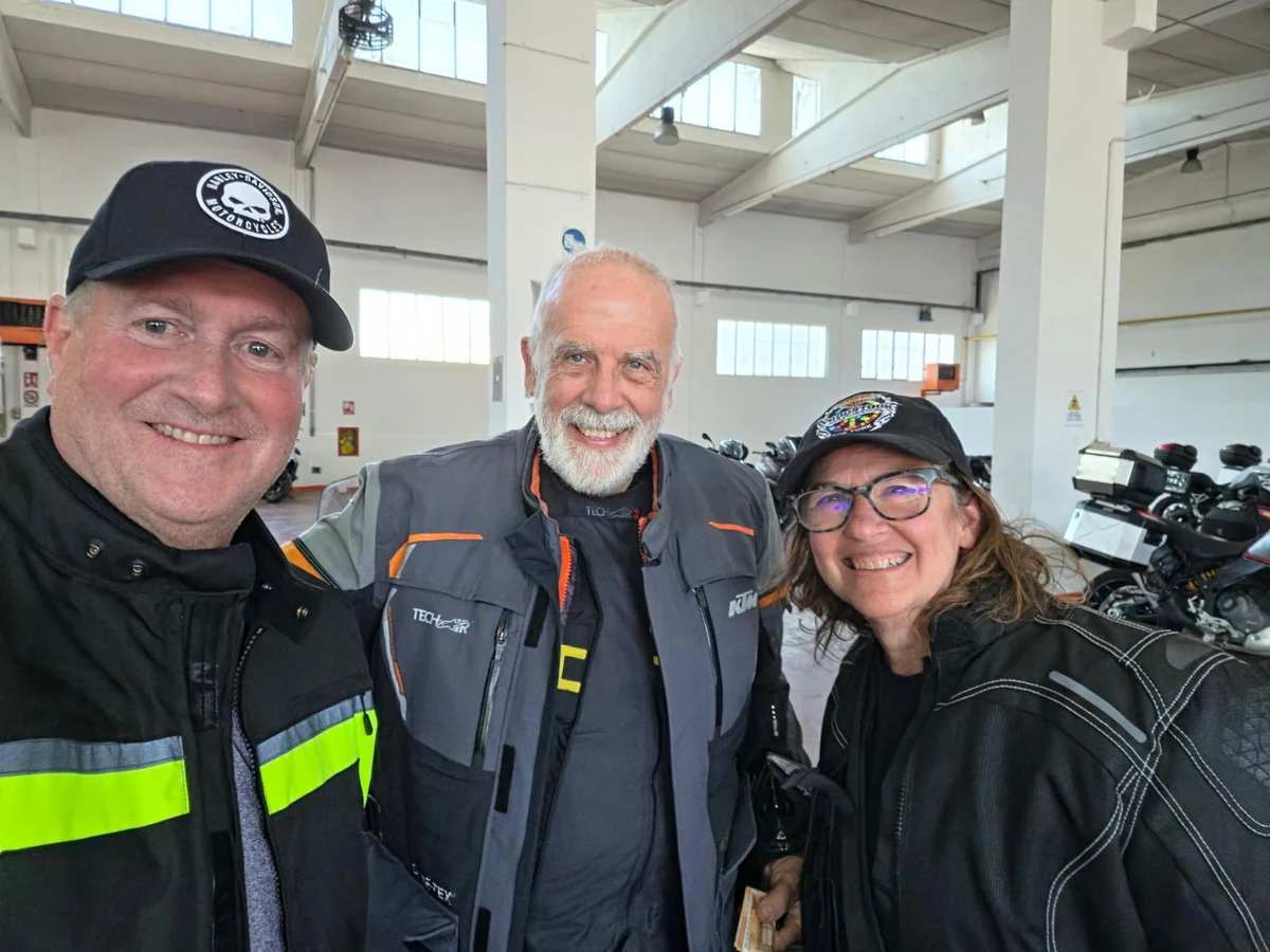 Three smiling motorcyclists in riding gear beside touring motorcycles inside a garage.