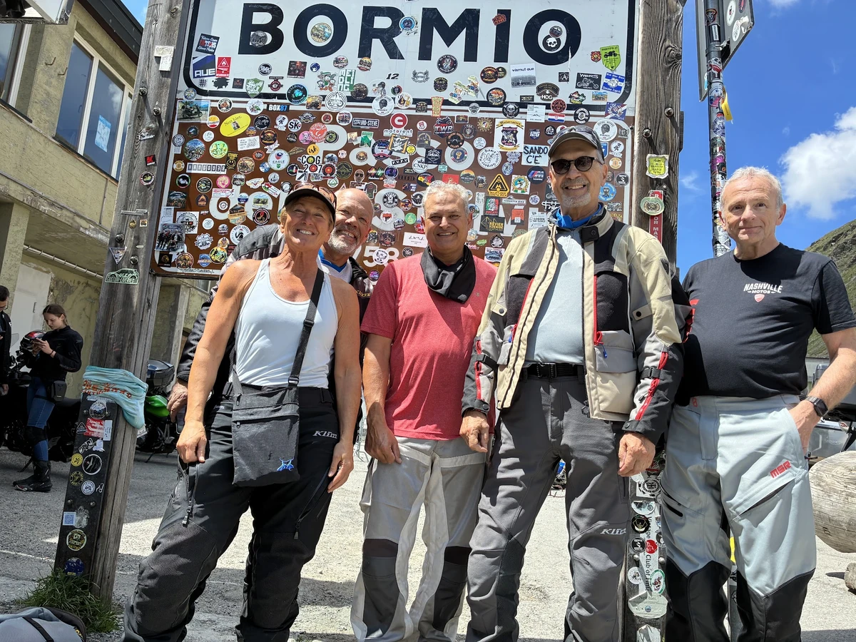 A group of motorcycle tour participants posing by the Bormio sign covered with travel stickers.