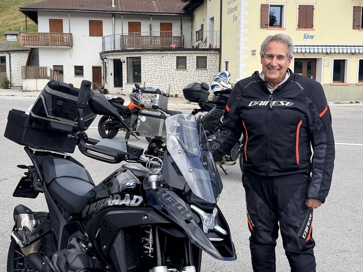 A smiling motorcycle tour participant beside a BMW touring motorcycle outside an alpine hotel.