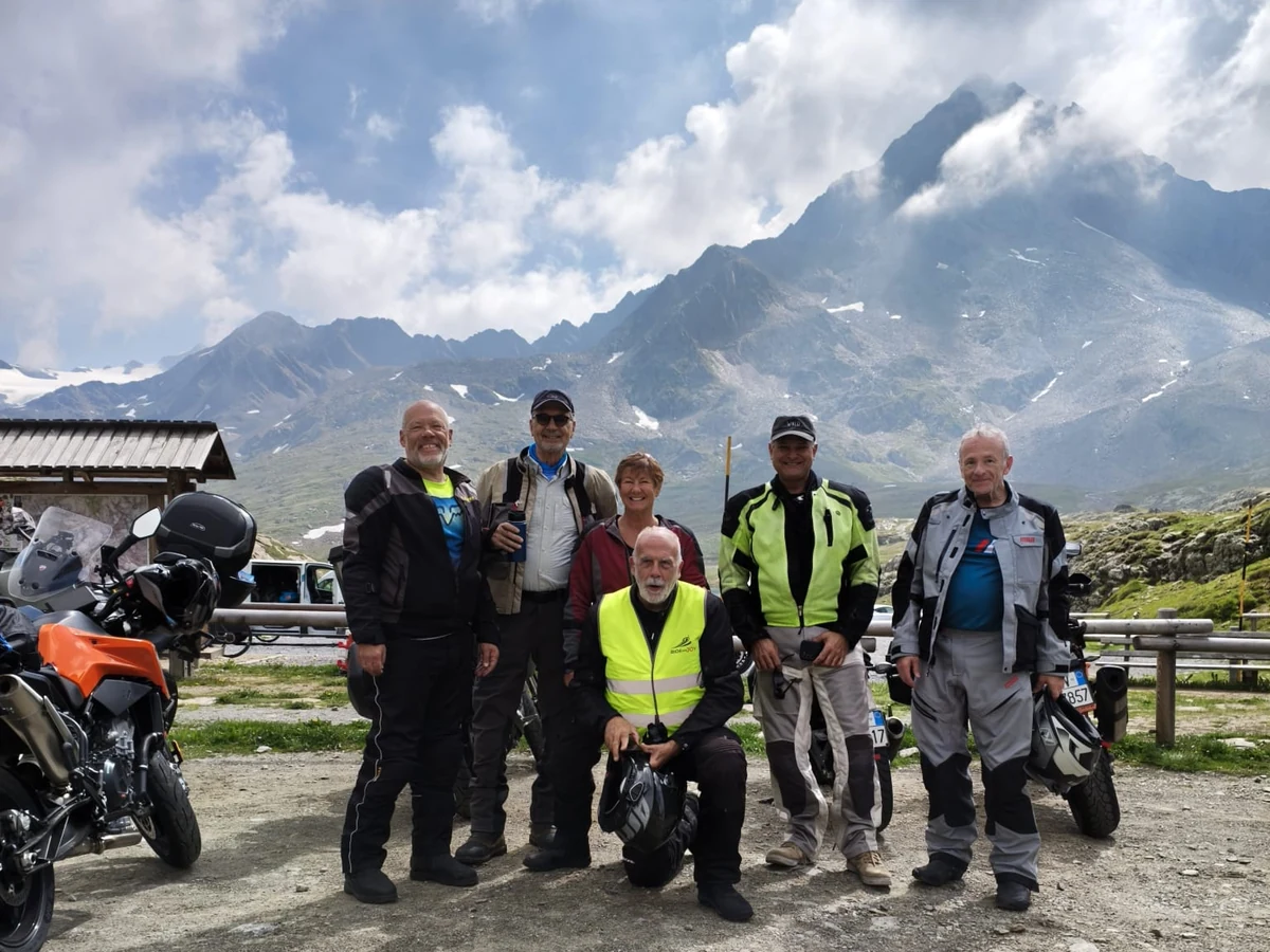 A group of motorcycle tour participants beside their bikes at a mountain pass, with rocky peaks behind them.