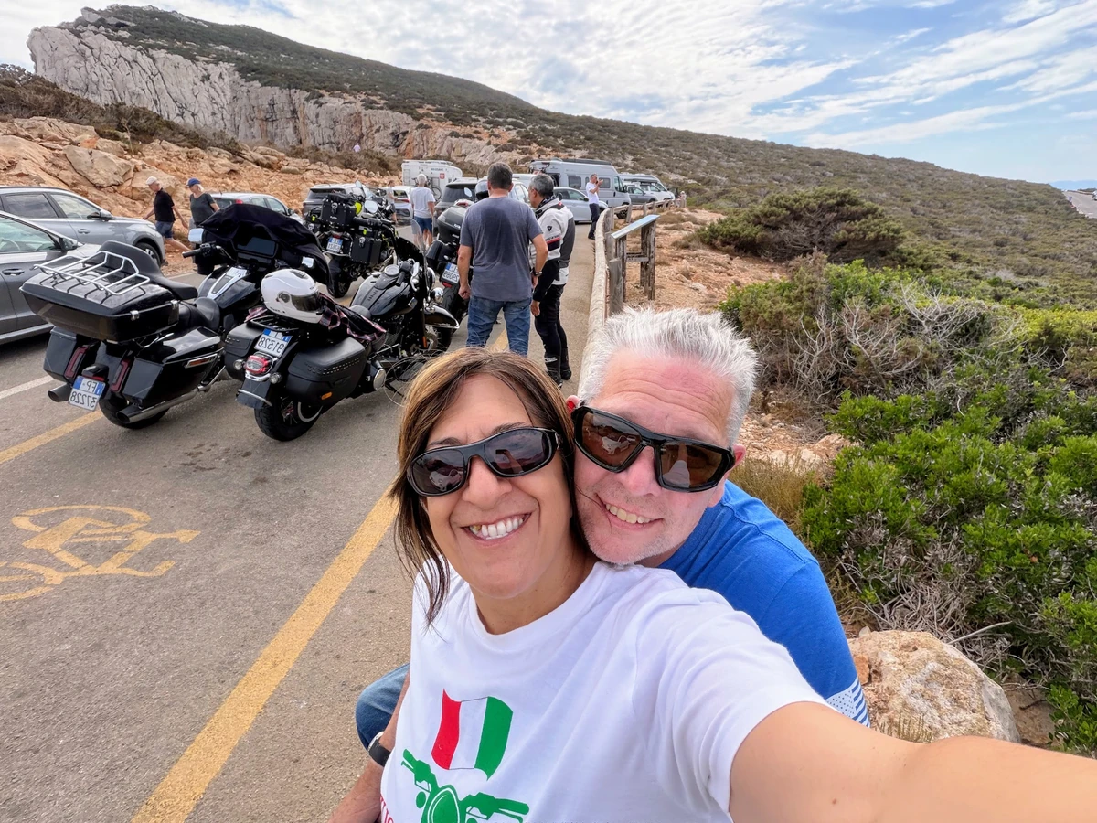 A smiling couple taking a selfie beside a coastal road, with touring motorcycles behind them.