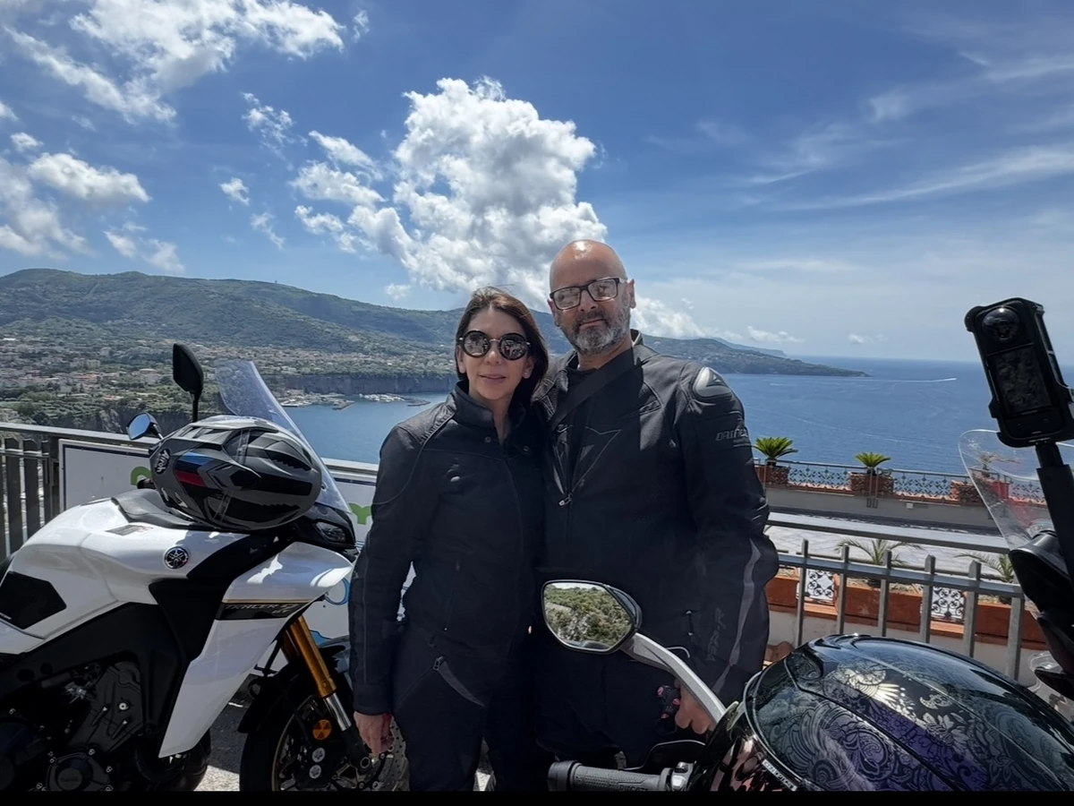 A couple in motorcycle riding gear beside motorcycles with the coast and sea behind them.