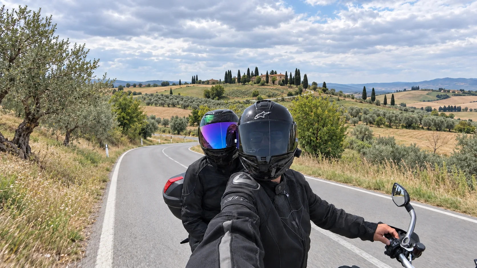 Two helmeted motorcycle travelers on a paved road through rolling countryside