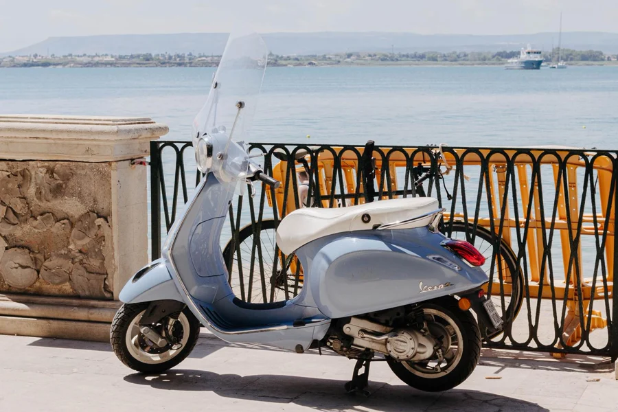 Light blue Vespa parked by a lakeside railing with bicycles locked behind it and boats on calm water in distance