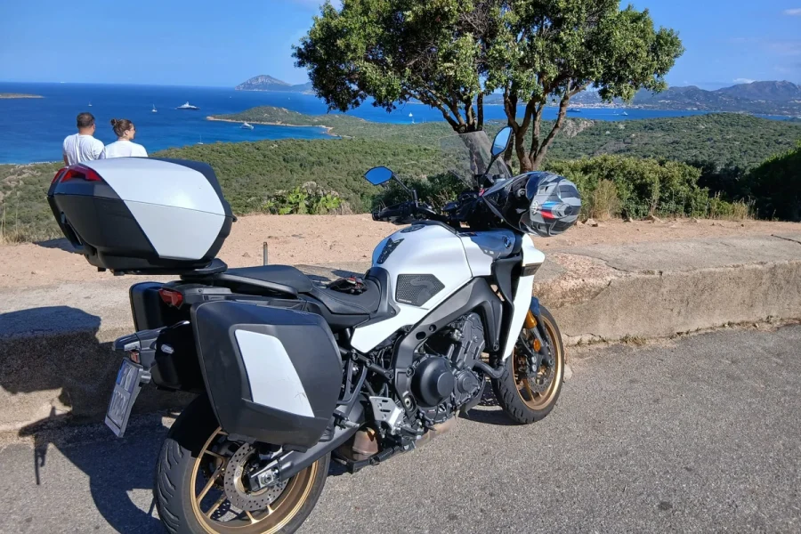 A touring motorcycle parked above the blue sea and islands along the Costa Smeralda coast in Sardinia.