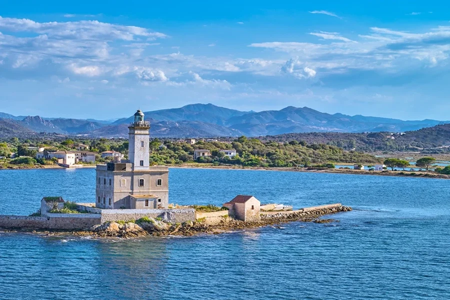 Historic lighthouse on a tiny islet, breakwater to Olbia’s gulf entrance; hills and coastal homes behind.