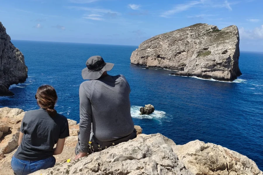 Two visitors looking out toward the Pan di Zucchero sea stack from the rocky coast at Masua, Sardinia.