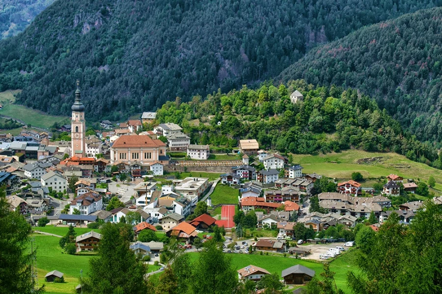 Tyrolean village with a tall church steeple, stone houses and green meadows at the base of forested mountains.